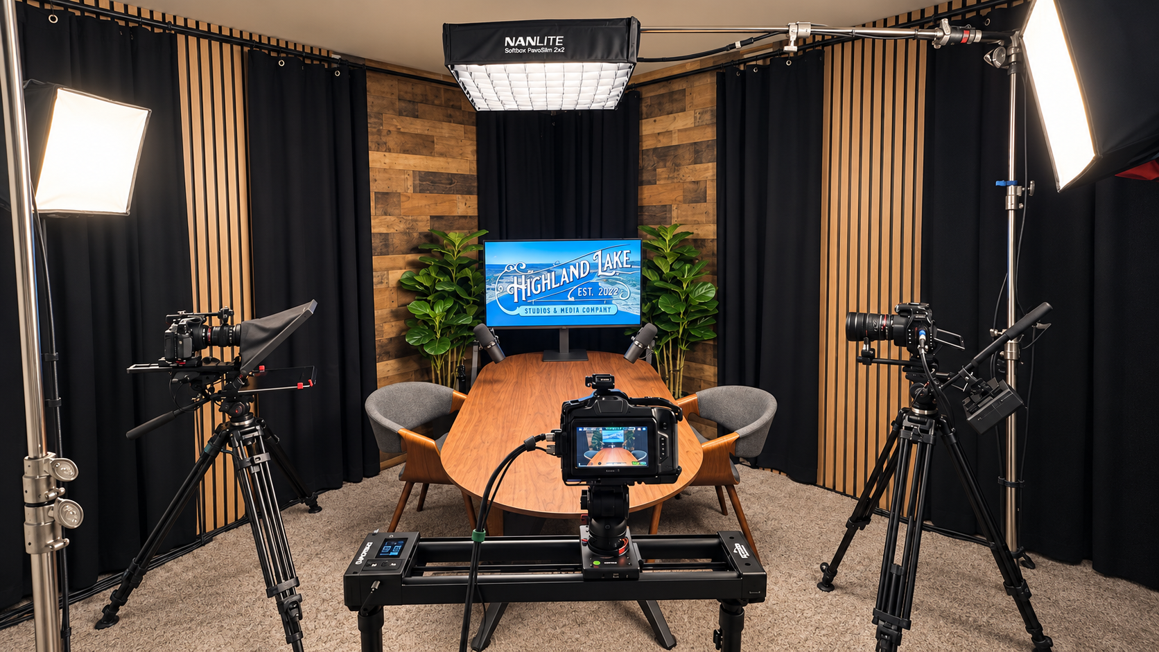 Highland Lake podcast studio with three cameras, a camera slider, two microphones, a wood table, and a monitor displaying the studio logo.